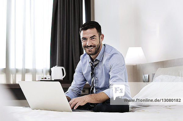 Portrait of happy businessman sitting with laptop on bed in hotel room