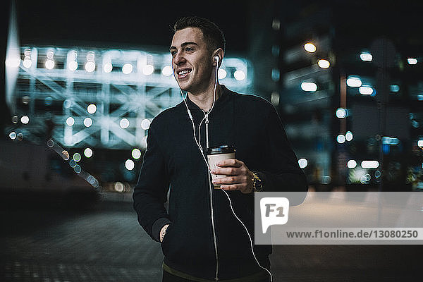 Lächelnder Mann mit Einwegbecher  der Musik hört  während er nachts gegen ein Gebäude in der Stadt steht