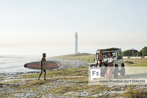 Mann mit Surfbrett geht am Strand bei klarem Himmel auf Freundinnen zu
