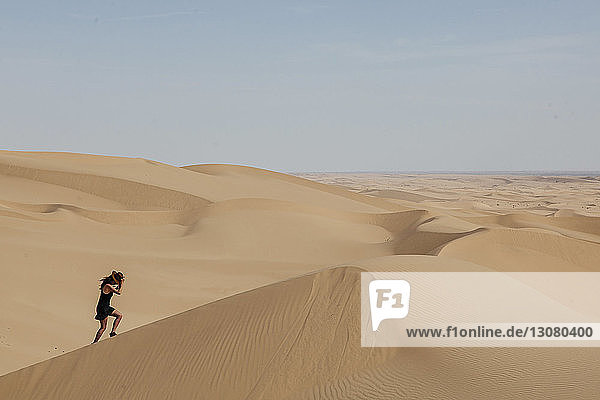 Seitenansicht einer jungen Frau mit Hut beim Klettern auf einer Sanddüne in der Wüste gegen den Himmel