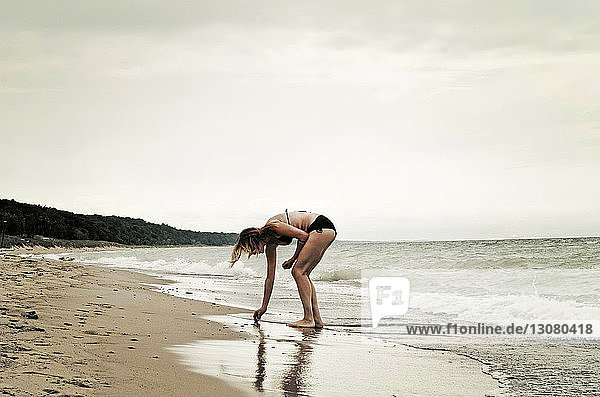 Frau im Bikini sammelt Muscheln am Strand vor klarem Himmel