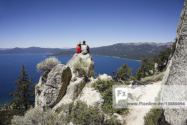Ehepaar sitzt auf einer Klippe mit Blick auf den See vor klarem blauen Himmel