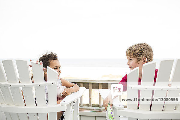 Happy siblings sitting on deck chairs at beach against clear sky