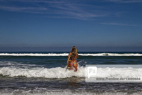 Rückansicht einer Frau  die ein Surfbrett trägt  während sie am sonnigen Tag im Meer vor blauem Himmel steht