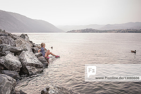 Side view of girl sitting by rocks at lakeshore against sky