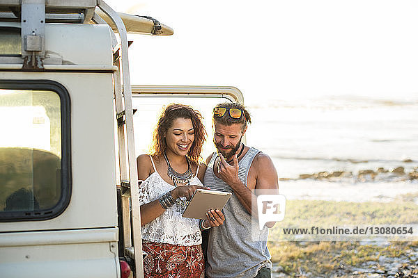 Frau zeigt ihrem Freund einen Tablet-Computer  während sie am Strand an einem Geländewagen steht