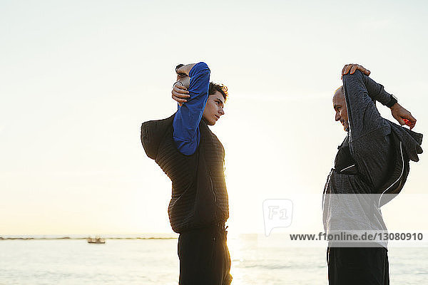 Side view of father and son stretching arms while standing face to face at beach against clear sky during sunset