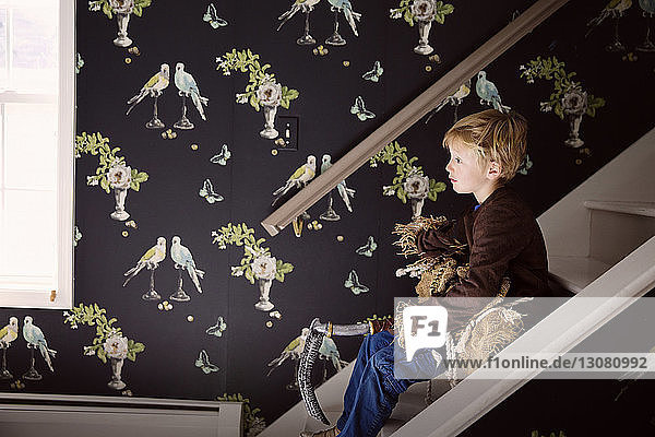 Seitenansicht eines Jungen  der ein Kostüm hält  während er zu Hause auf einer Treppe sitzt