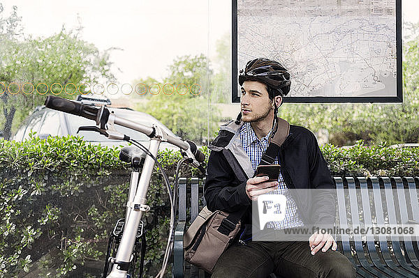 Junger männlicher Pendler hält Smartphone in der Hand  während er an der Bushaltestelle sitzt