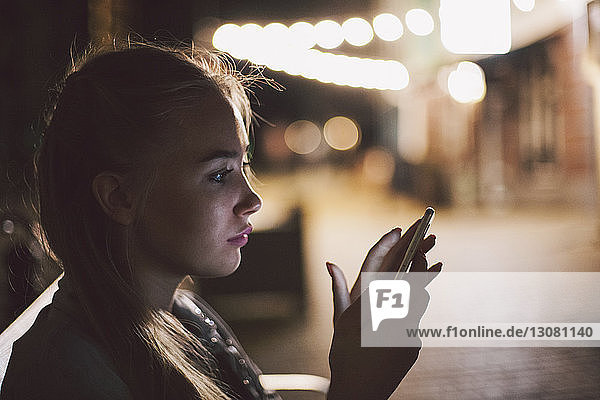 Junge Frau benutzt Smartphone auf beleuchtetem Bürgersteig in der Stadt während der Nacht