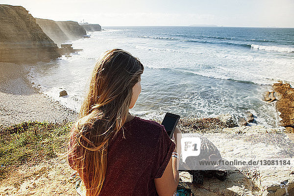 Rückansicht einer Frau  die ein Mobiltelefon benutzt  während sie auf einer Klippe am Meer sitzt