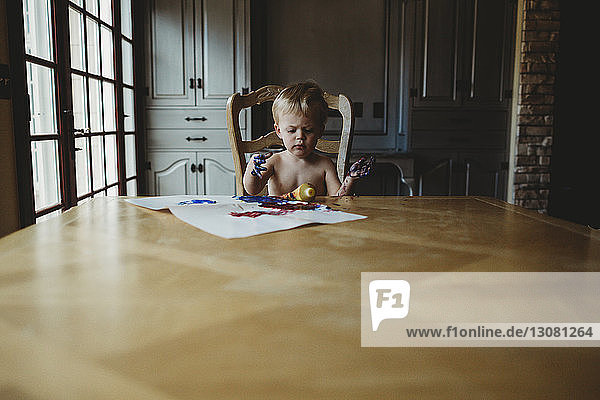 Kleiner Junge ohne Hemd mit schmutzigen Händen  der zu Hause am Holztisch sitzt
