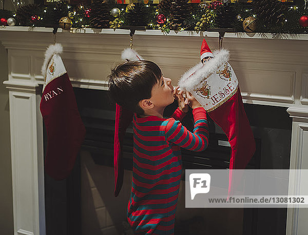 Seitenansicht eines neugierigen Jungen  der ein Spielzeug in Socken betrachtet  das zu Weihnachten am Kamin hängt