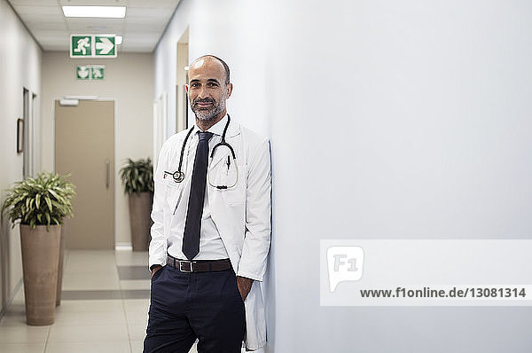 Porträt eines lächelnden Arztes mit an die Wand gelehnten Händen in Taschen im Krankenhaus