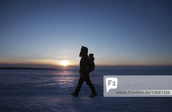 Side view of woman walking on snow covered field against sky during sunset