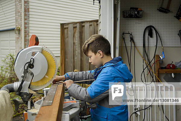 Side view of boy measuring wooden plank while working at backyard