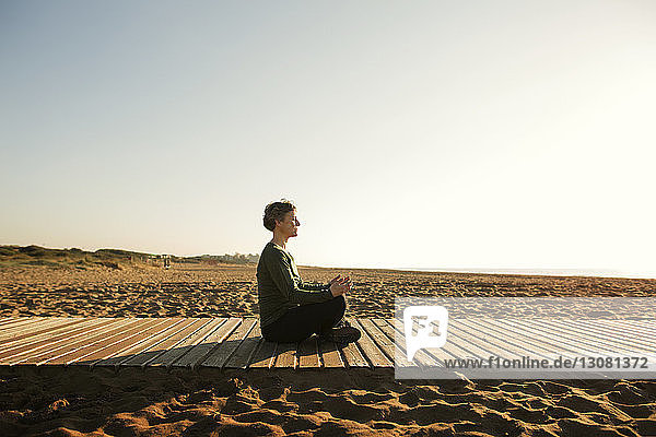 Side view of woman meditating on footpath at beach against clear sky