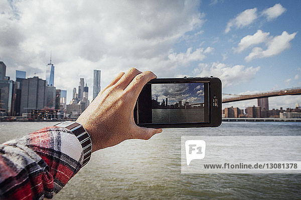 Cropped hand of man photographing skyline and Brooklyn Bridge