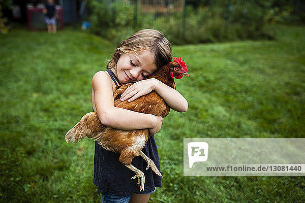 Smiling girl with eyes closed holding hen while standing in yard