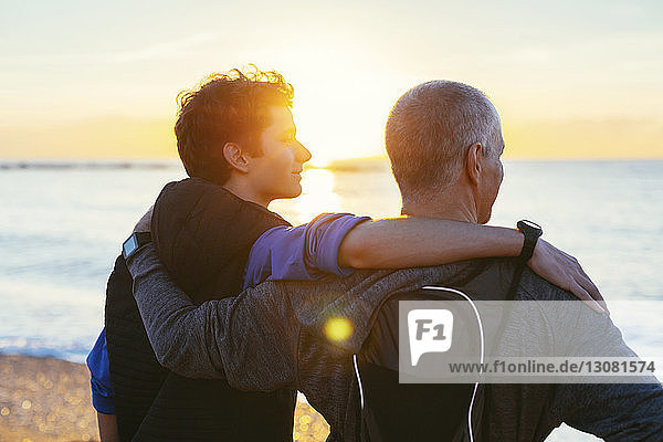 Vater und Sohn mit Armen um die Schultern  die am Strand stehen und auf die Aussicht schauen