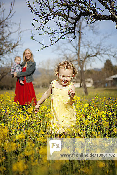 Fröhliches Mädchen rennt auf dem Feld  während die Mutter mit dem Kleinkind im Hintergrund steht