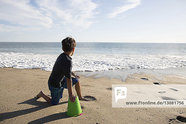 Boy looking at sea while sitting with bucket on beach during sunny day