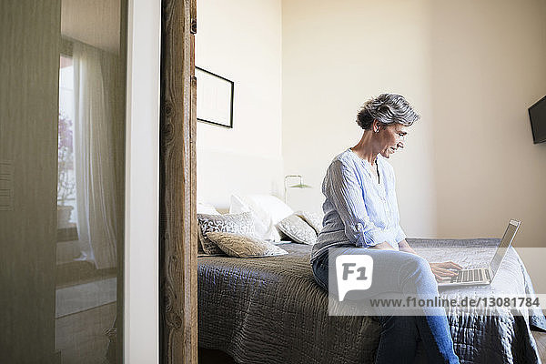Side view of mature woman using laptop while sitting on bed at home