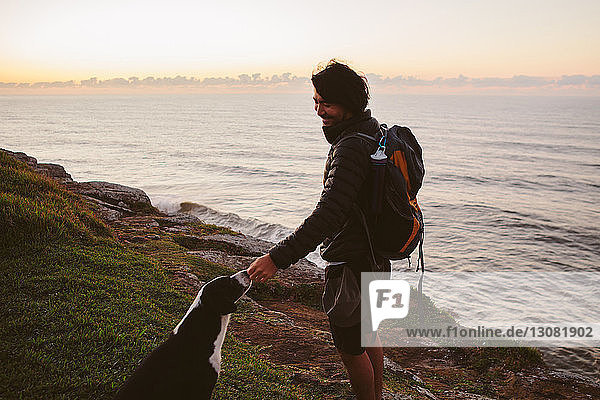 Side view of male hiker touching dog while standing on hill by sea