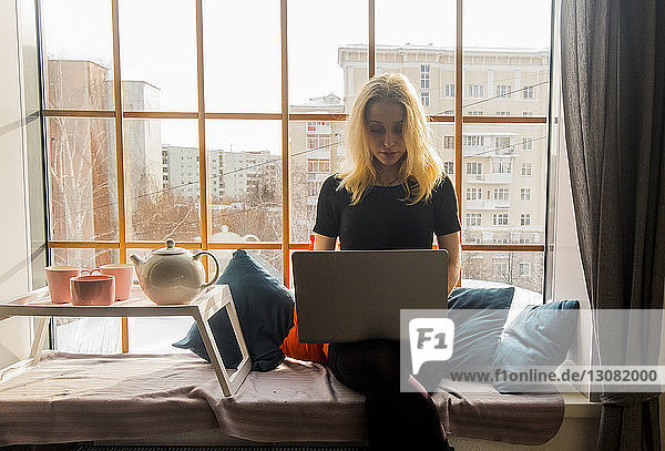 Frau benutzt Laptop-Computer auf einem Hocker am Fensterplatz in einer Nische zu Hause