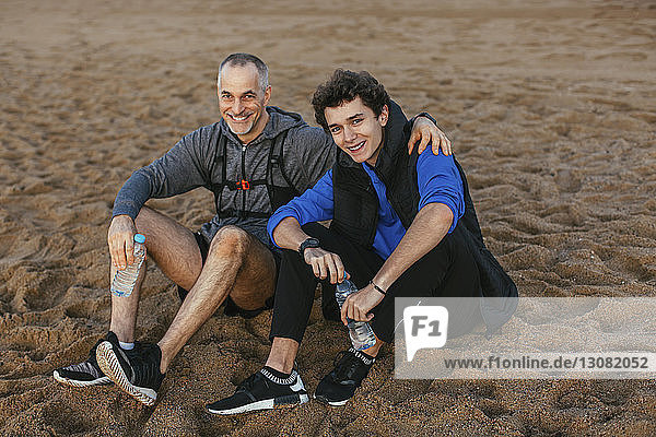 High angle portrait of happy father and son holding water bottles while sitting at beach