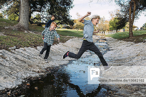 Brothers jumping over stream at park