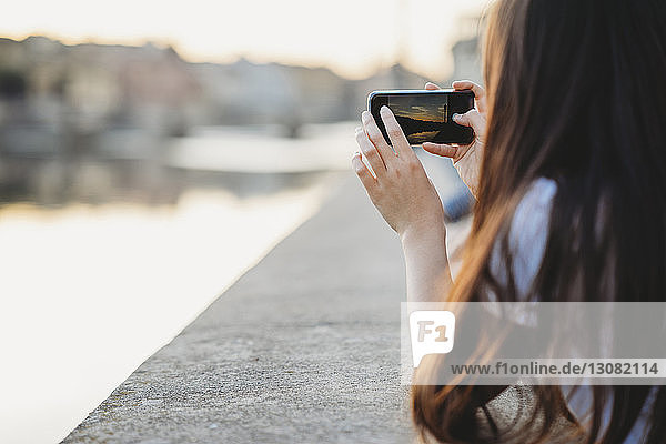 Seitenansicht einer Frau  die bei Sonnenuntergang mit einem Mobiltelefon an einer Stützmauer in der Stadt fotografiert