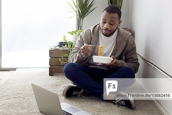 Geschäftsmann isst Nudeln  während er im Kreativbüro auf den Laptop am Boden schaut