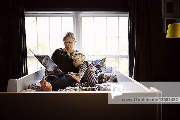 Mother reading book to curious son while sitting on bed against window at home
