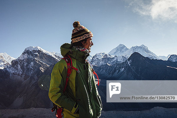 Wanderer in warmer Kleidung schaut weg  während er auf einem Berg vor blauem Himmel steht
