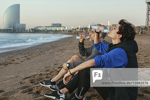 Father and son drinking water while sitting against Hotel Vela at beach
