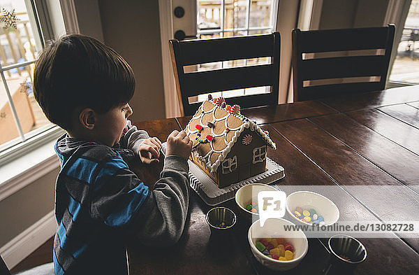 Schrägaufnahme eines Jungen  der zu Weihnachten zu Hause Lebkuchenhaus auf einem Holztisch backt