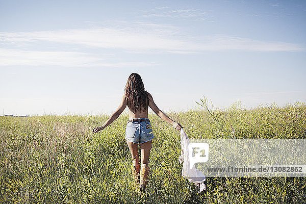 Rear view of woman holding shirt and walking on grassy field against sky