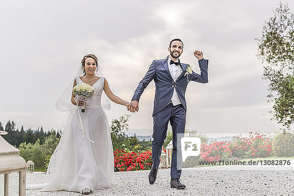 Glückliches frisch verheiratetes Paar hält sich an den Händen  während es auf einem Fußweg gegen den bewölkten Himmel geht