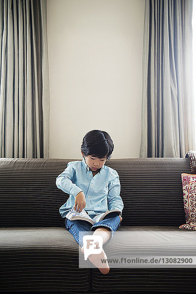 Boy reading book while sitting on sofa against wall at home