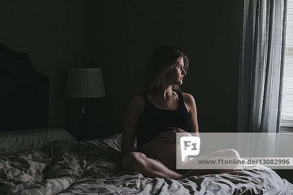 Schwangere Frau schaut weg  während sie zu Hause auf dem Bett sitzt