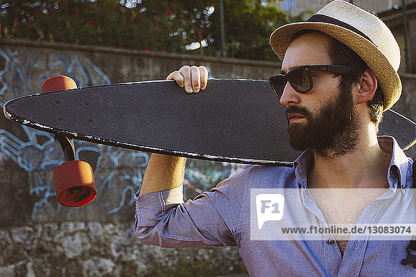 Mann mit Sonnenbrille und Hut  während er ein Skateboard an die Wand hält