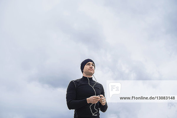 Low angle view of thoughtful male athlete listening music against cloudy sky