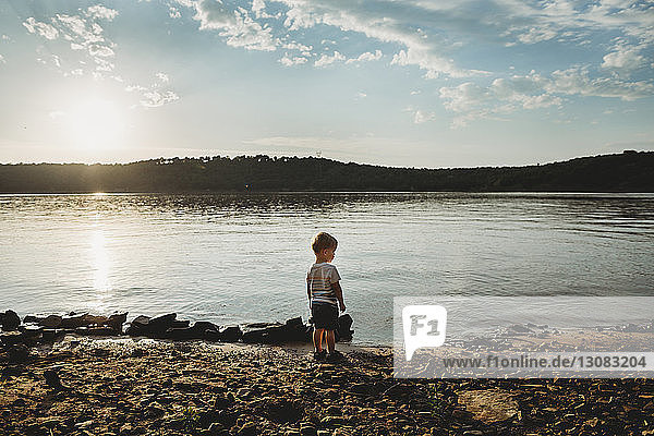 Rückansicht eines kleinen Jungen  der bei Sonnenuntergang am Seeufer gegen den Himmel steht