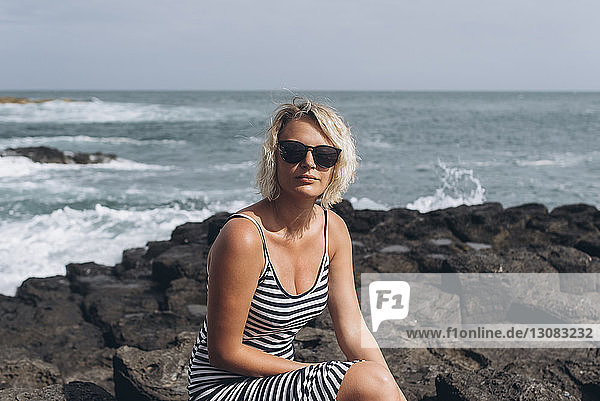 Portrait of confident woman wearing sunglasses while sitting on rocks at beach against sea