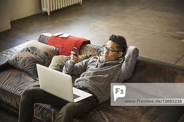 High angle view of businessman with laptop computer using phone while reclining on sofa in creative office