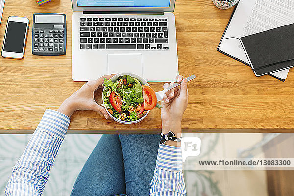 Overhead view of woman holding salad bowl by laptop computer at wooden table