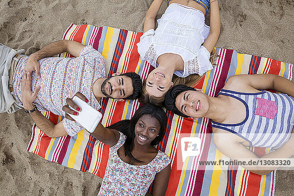 Overhead view of multi-ethnic friends taking selfie while lying on huddle on beach