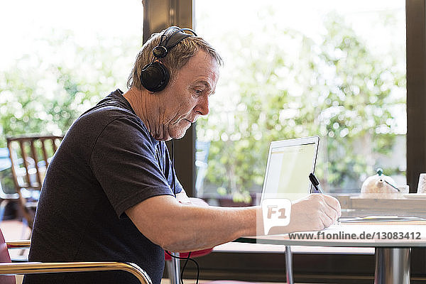 Seitenansicht eines älteren Mannes  der schreibt  während er zu Hause über seinen Laptop Musik hört