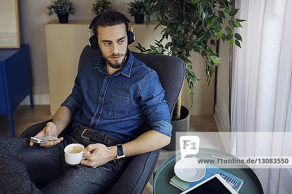 Man holding coffee cup and mobile phone listening music while sitting on chair at home